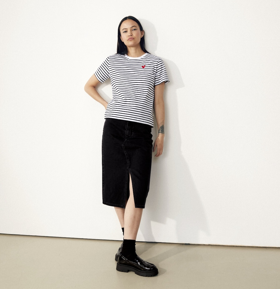Eine Frau lehnt sich an die Wand an und trägt ein tshirt mit Streifen und einen schwarzen Rock, der über das Knie geht.