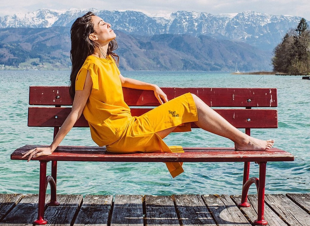 Eine Frau sitzt auf einer Bank am See und geniesst die Sonne mit Kleidern von Anukoo.